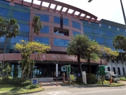 A long seven story building with red and white accents on a cloudy day. There are at least six cars in the parking lot in front of the building.